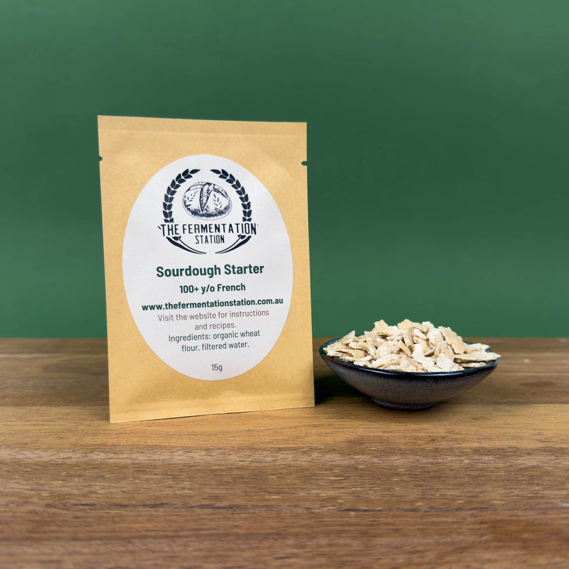 Sourdough starter retail pouch from The Fermentation Station displayed on a wooden surface beside a small blue bowl showing the dried starter flakes, with a simple green background.
