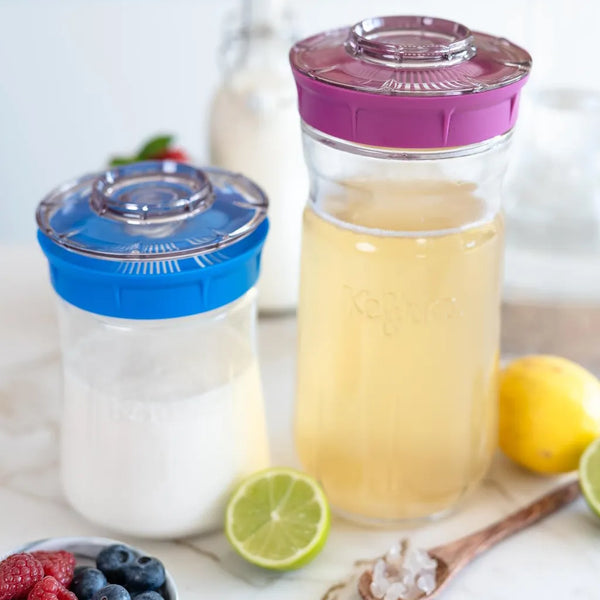 Kefirko kefir fermenters with blue and purple lids on kitchen counter, filled with milk kefir and water kefir alongside fresh fruit and lime