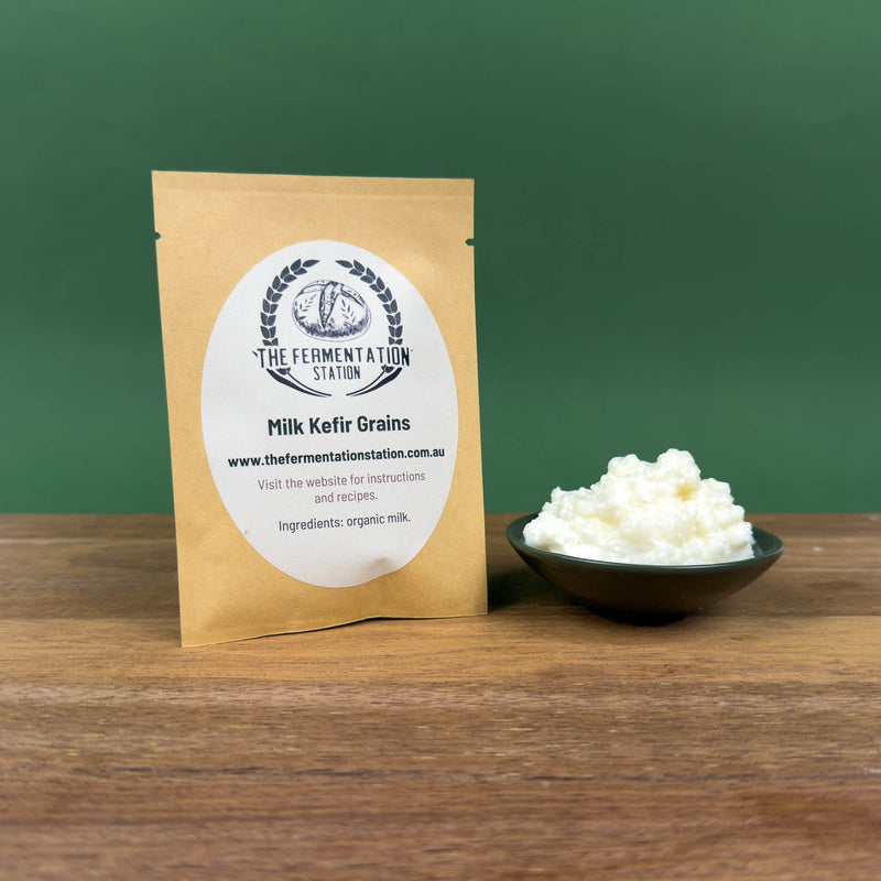 Milk Kefir grains retail pouch from The Fermentation Station displayed on a wooden surface beside a small green bowl showing the milk kefir grains, with a simple green background.