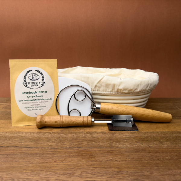 sourdough baking kit containing sourdough starter, banneton, lame, danish dough whisk and bench scraper on wooden board