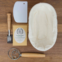 sourdough baking kit containing sourdough starter, banneton, lame, danish dough whisk and bench scraper flat lay on wooden board