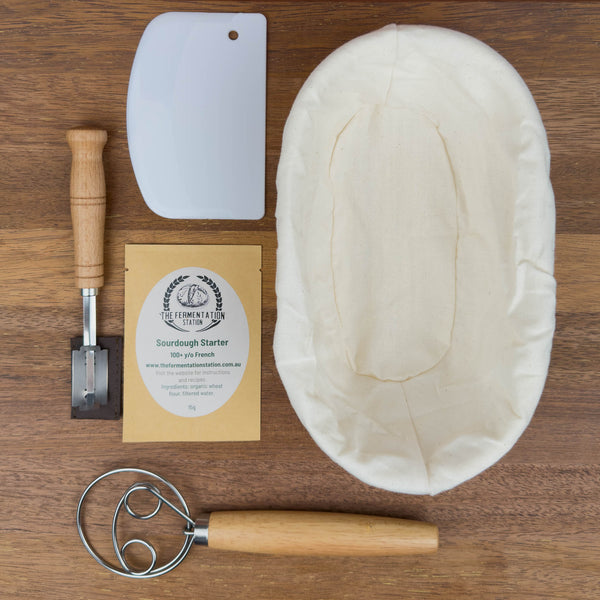 sourdough baking kit containing sourdough starter, banneton, lame, danish dough whisk and bench scraper flat lay on wooden board