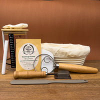 sourdough master kit containing sourdough starter, banneton, lame, danish dough whisk, bench scraper, glass jar, spatula and labels on wooden board
