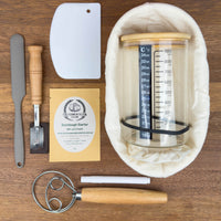 sourdough master kit containing sourdough starter, banneton, lame, danish dough whisk, bench scraper, glass jar, spatula and labels flat lay on wooden board