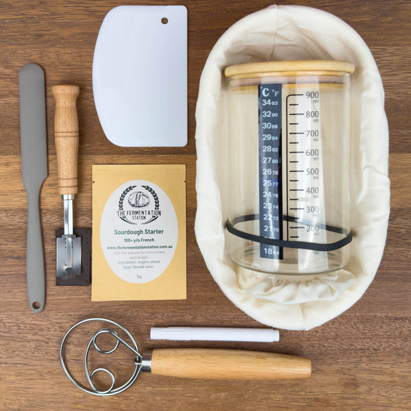 sourdough master kit containing sourdough starter, banneton, lame, danish dough whisk, bench scraper, glass jar, spatula and labels flat lay on wooden board