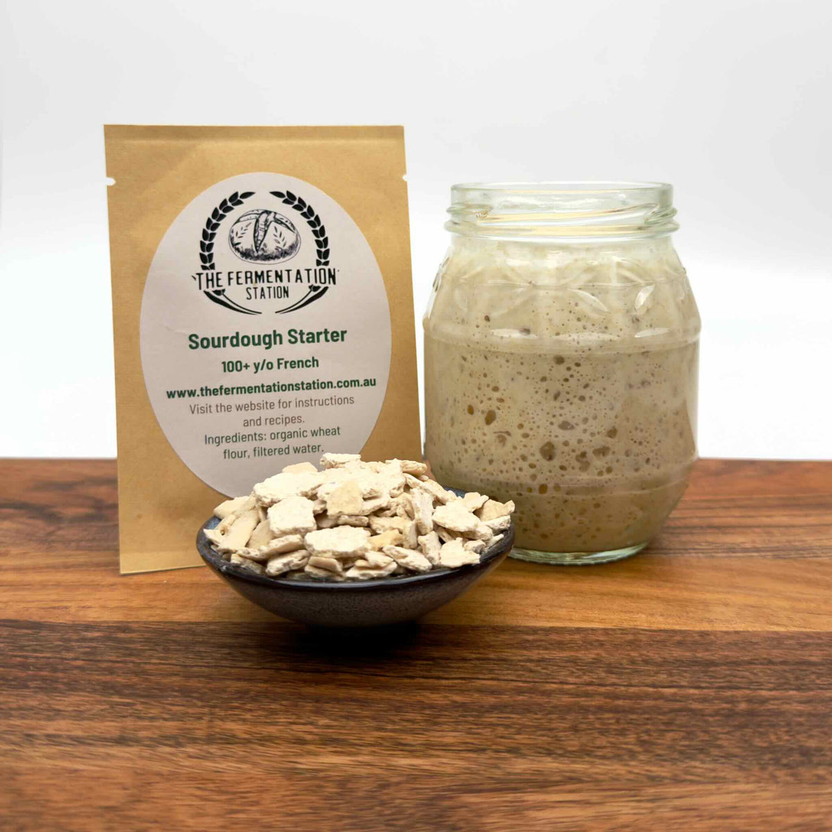 Sourdough starter, package, bubbling starter in a jar, and bowl on a wooden surface with 'The Fermentation Station' branding.