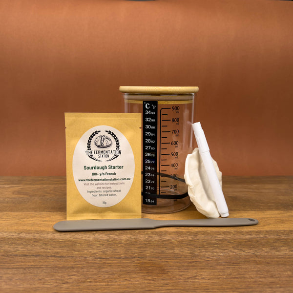 sourdough starter kit containing sourdough starter, glass jar with wood and linen lids, silicone spatula, pen and labels on wooden board
